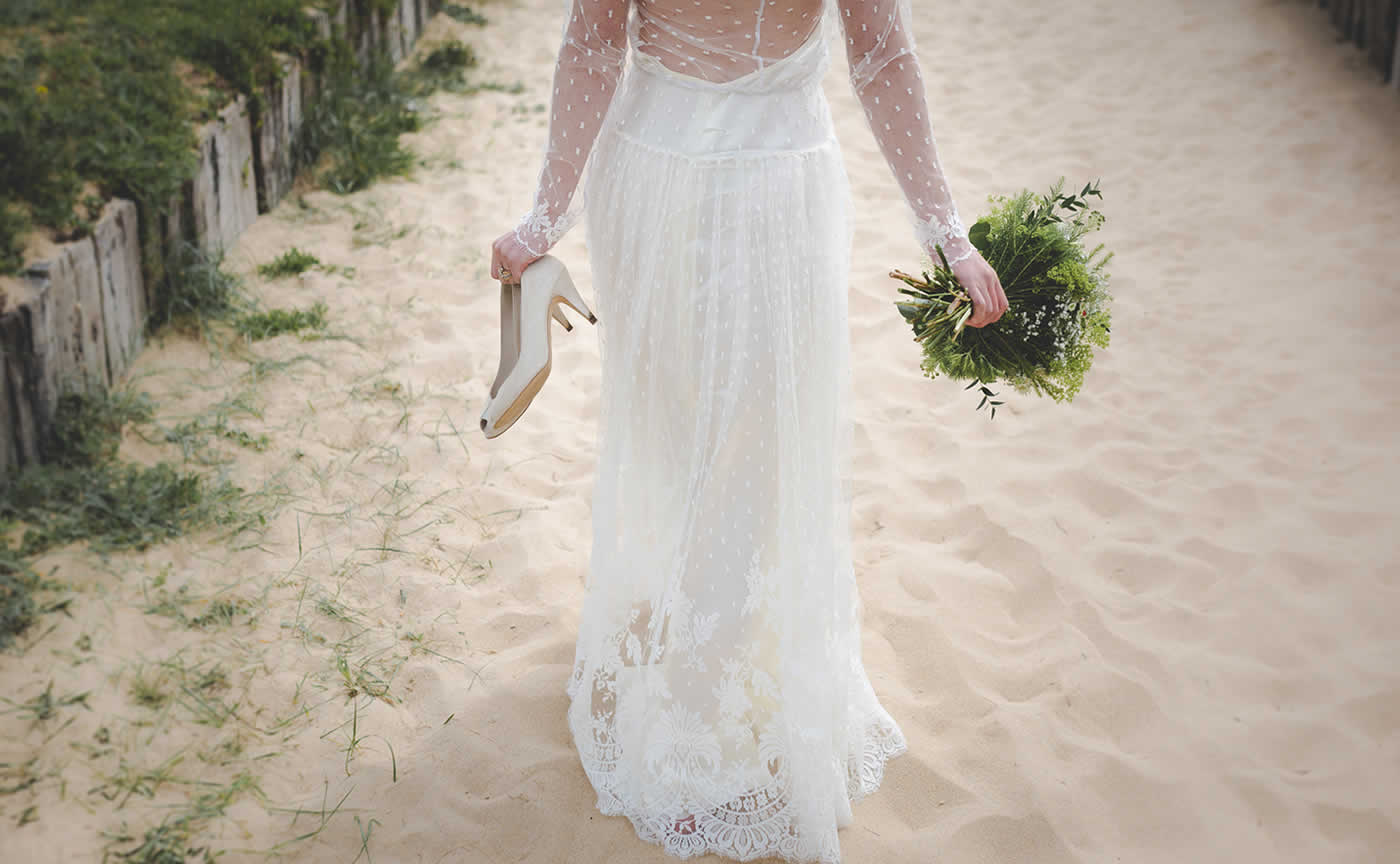 ハイヒールと緑のブーケを手に砂浜の上を歩く白ドレス姿の花嫁